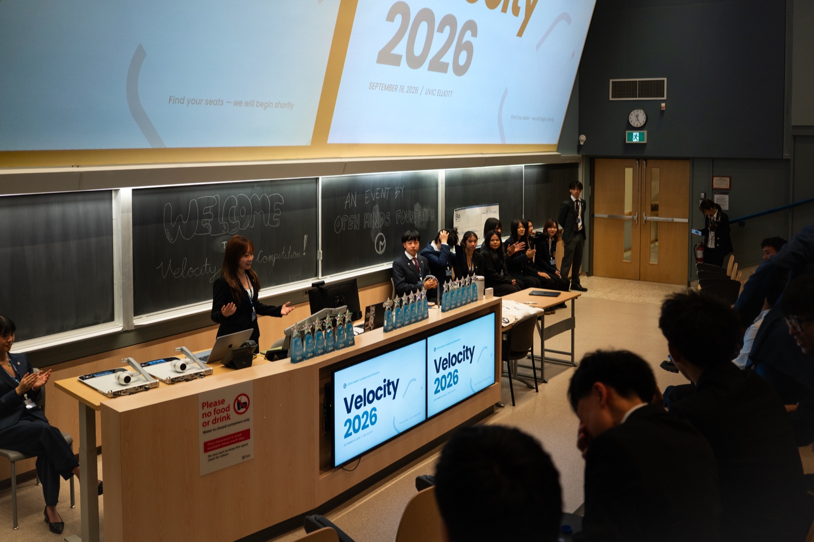 Velocity 2026 在 University of Victoria 讲堂举行开幕致辞;筹办团队站在前方,桌上摆放奖杯,黑板上写着欢迎参加 Velocity Case Competition。